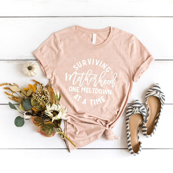 May include a white unisex graphic T-shirt laid flat with the words “Surviving Motherhood One Meltdown at a Time” printed in bold lettering across the chest.
