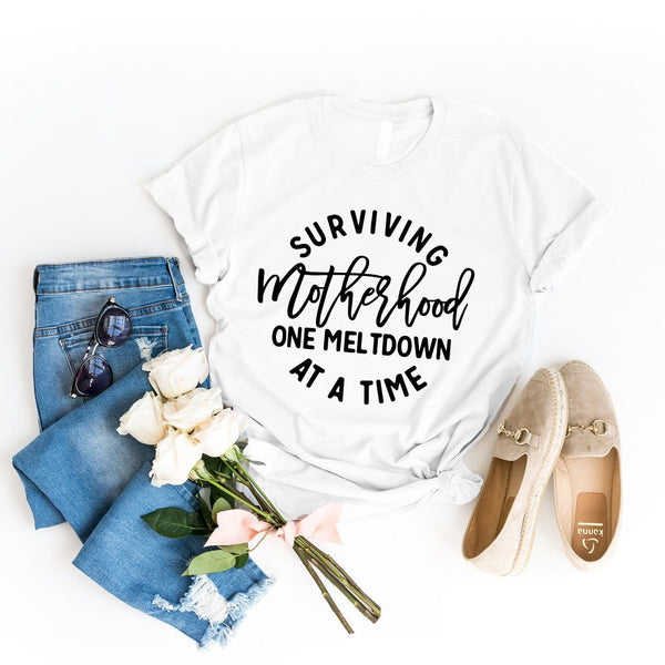 May include a white unisex graphic T-shirt laid flat with the words “Surviving Motherhood One Meltdown at a Time” printed in bold lettering across the chest.