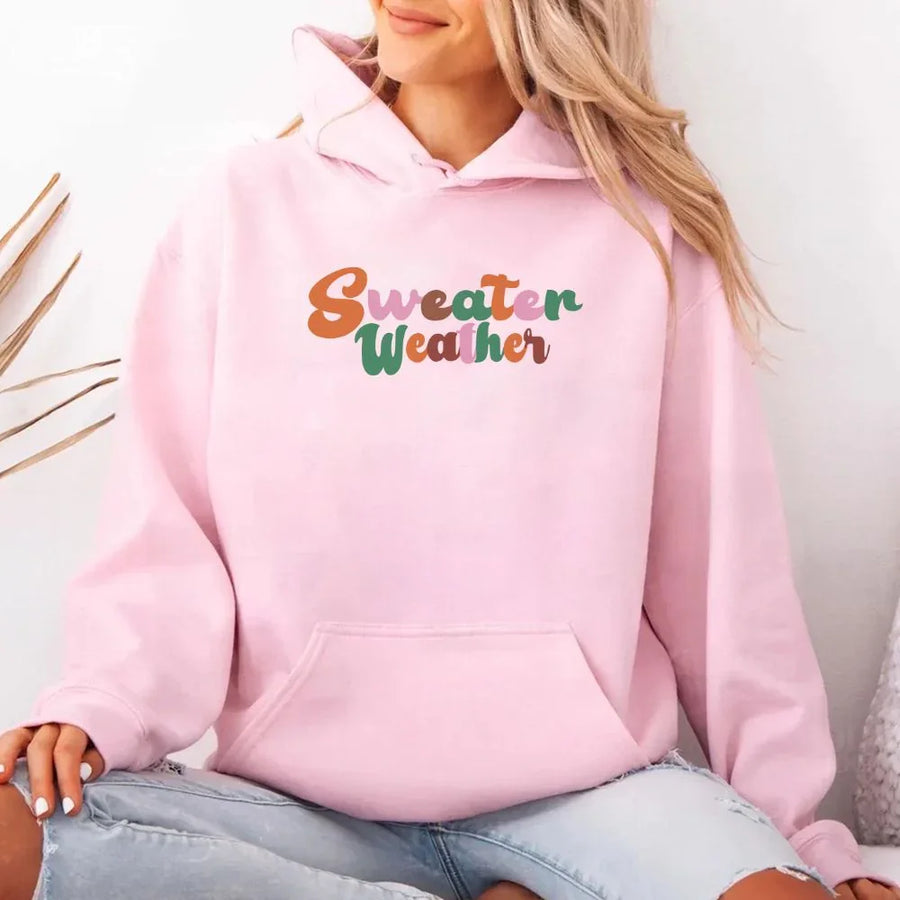 Person wearing a pink hoodie with 'Sweater Weather' text, sitting on a white background.