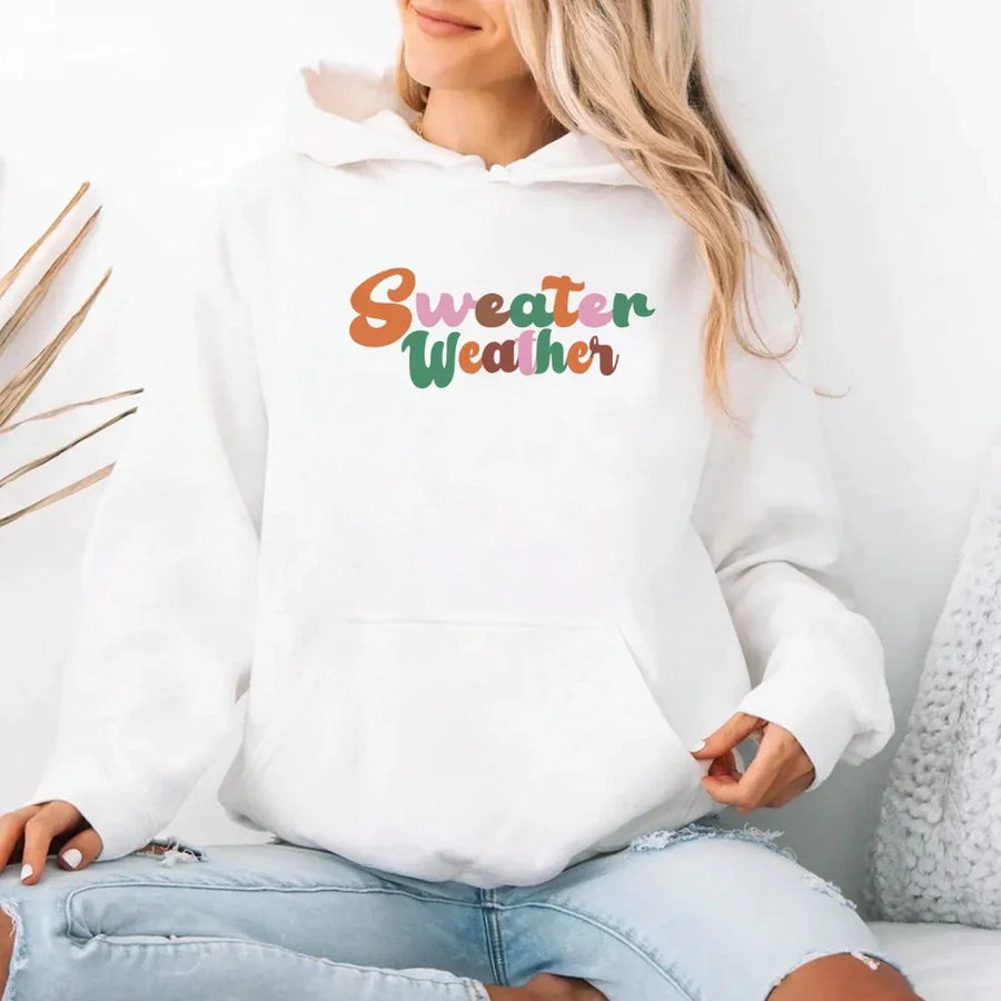 White hoodie with 'Sweater Weather' text worn by a person sitting on a white couch.