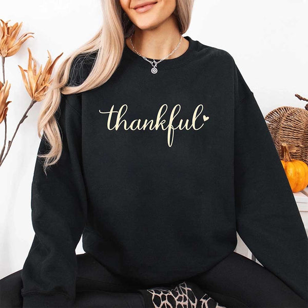 Wearing a black sweatshirt with the design 'thankful' for Thanksgiving, sitting in a cozy room with pumpkins and decorative elements.