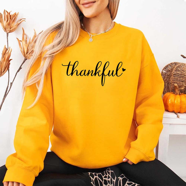 Wearing a gold sweatshirt with the design 'thankful' for Thanksgiving, sitting in a cozy room with pumpkins and decorative elements.
