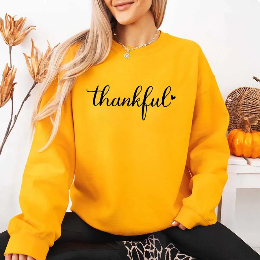 Wearing a gold sweatshirt with the design 'thankful' for Thanksgiving, sitting in a cozy room with pumpkins and decorative elements.