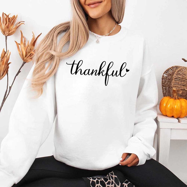 Wearing a white sweatshirt with the design 'thankful' for Thanksgiving, sitting in a cozy room with pumpkins and decorative elements.
