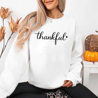 Wearing a white sweatshirt with the design 'thankful' for Thanksgiving, sitting in a cozy room with pumpkins and decorative elements.