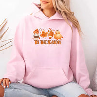 Person wearing a pink hoodie with 'Tis the Season' text and autumn graphics.