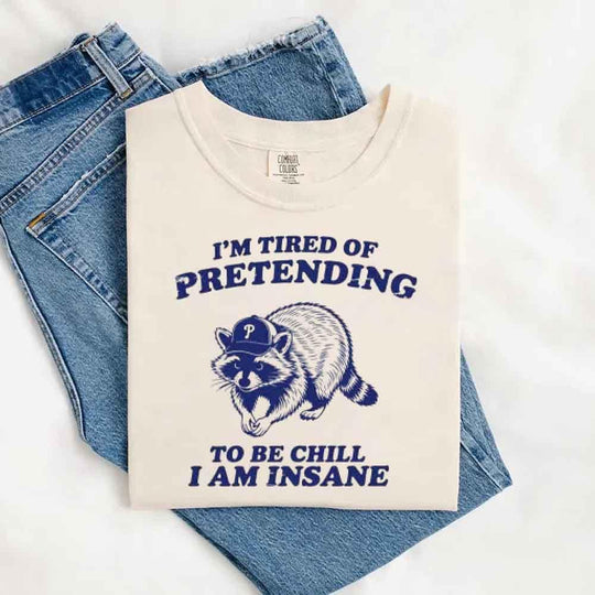 May include a woman sitting on a bed wearing an Ivory Comfort Colors T-shirt that says “I’m tired of pretending to be chill I am insane” with a cartoon raccoon wearing a cap.