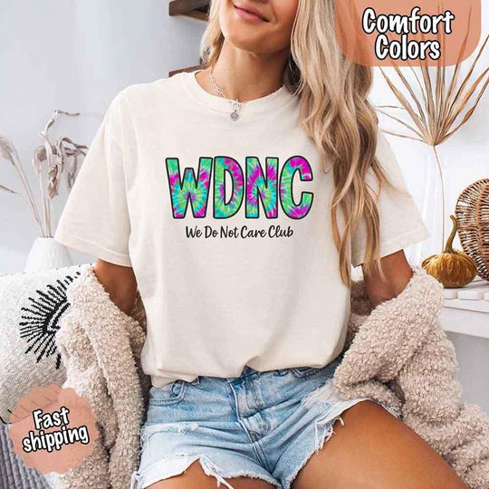 May include a woman wearing a ivory Comfort Colors t-shirt with 'WDNC – We Do Not Care Club' tie-dye text, seated with cozy cardigan and denim shorts. Fast shipping badge included.
