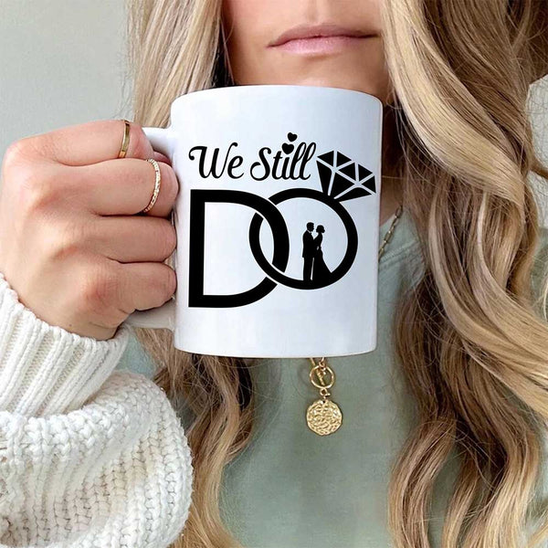 Person holding a white mug with 'We Still Do' design featuring wedding rings and a couple.