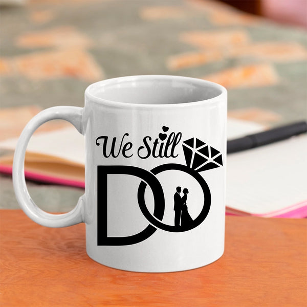 White mug with 'We Still Do' design featuring a couple and diamond, on a wooden surface.