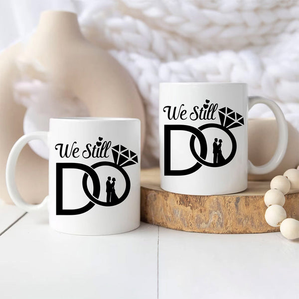 Two white mugs with 'We Still Do' design on a wooden surface.