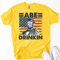Yellow t-shirt with patriotic Abraham Lincoln, American flag, and 'Abe Drinkin' text for July 4th
