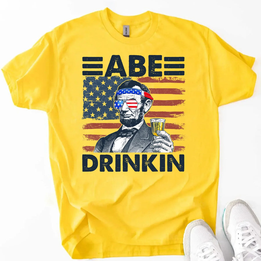 Yellow t-shirt with patriotic Abraham Lincoln, American flag, and 'Abe Drinkin' text for July 4th