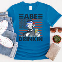 Blue 4th of July shirt with Abraham Lincoln holding a drink, USA flag background, patriotic tee