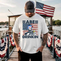 Man wearing a 4th of July Abe Drinkin patriotic shirt with American flag at a festive dock.