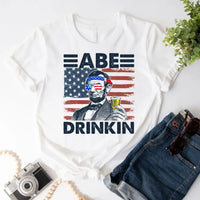 White 4th of July shirt with Abraham Lincoln holding a drink, American flag background, denim shorts.