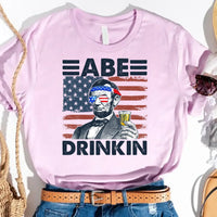 Pink 4th of July t-shirt featuring Abraham Lincoln with beer, USA flag sunglasses, and "Abe Drinkin" text.