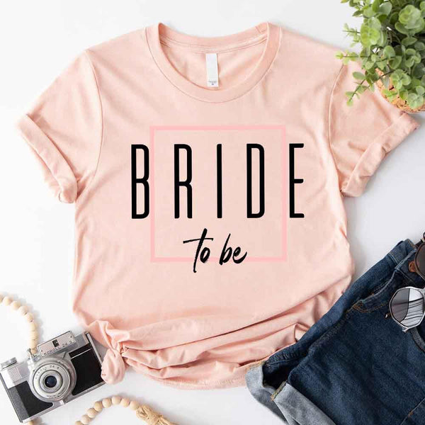 Peach t-shirt with 'BRIDE to be' text on a white surface with jeans and a camera.
