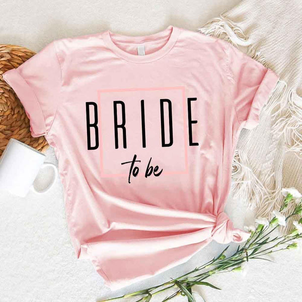 Pink t-shirt with 'BRIDE to be' text on a white surface with flowers and a woven basket.