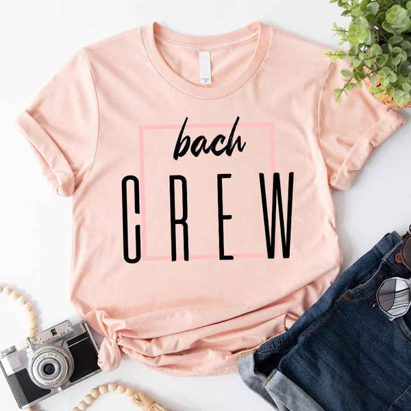 Peach T-shirt with 'bach CREW' text on a white surface with jeans and camera.
