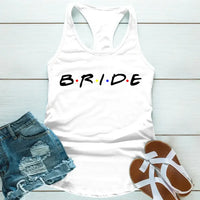 White tank top with the word 'BRIDE' printed in black letters, positioned on a wooden surface with blue denim shorts and white sandals in the background.