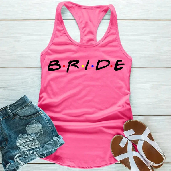 Pink tank top with the word 'BRIDE' printed in black letters, positioned on a wooden surface with blue denim shorts and white sandals in the background.