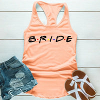 Peach tank top with the word 'BRIDE' printed in black letters, positioned on a wooden surface with blue denim shorts and white sandals in the background.
