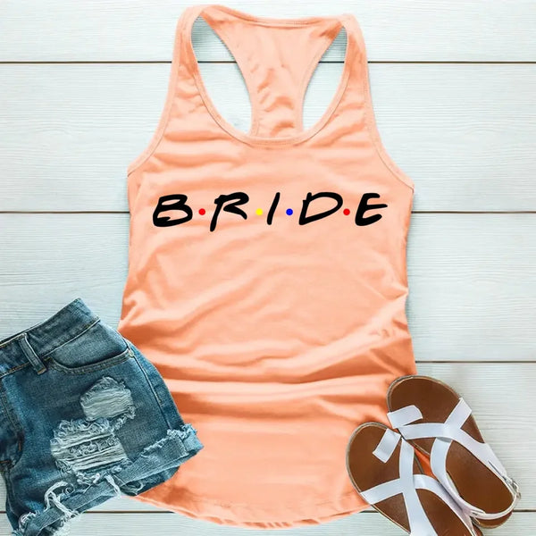 Peach tank top with the word 'BRIDE' printed in black letters, positioned on a wooden surface with blue denim shorts and white sandals in the background.