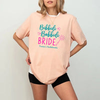May include Peach a personalized Bibbidi Bobbidi Bride T-shirt with fairytale bridal lettering for a bachelorette party or wedding celebration.