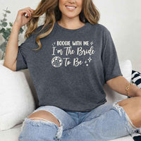 May include a Dark Heather retro-style graphic tee with bold disco lettering reading “Boogie With Me, I’m The Bride To Be,” featuring playful fonts and festive design elements!