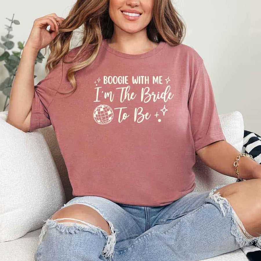 May include a Mauve retro-style graphic tee with bold disco lettering reading “Boogie With Me, I’m The Bride To Be,” featuring playful fonts and festive design elements!