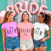 Three women wearing 'Bride to be' and 'Beach Crew' shirts with pink and silver balloons in the background.