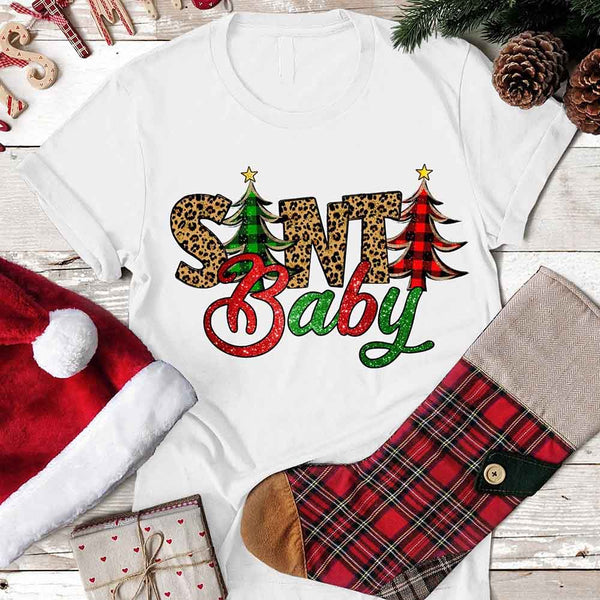 White t-shirt with 'Santa Baby' design on a wooden surface with Christmas decorations.