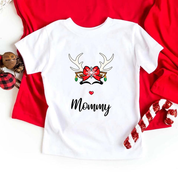 Folded red or white unisex Christmas T-shirt with a simple reindeer graphic design, styled beside ornaments and pine décor for a cozy holiday look.