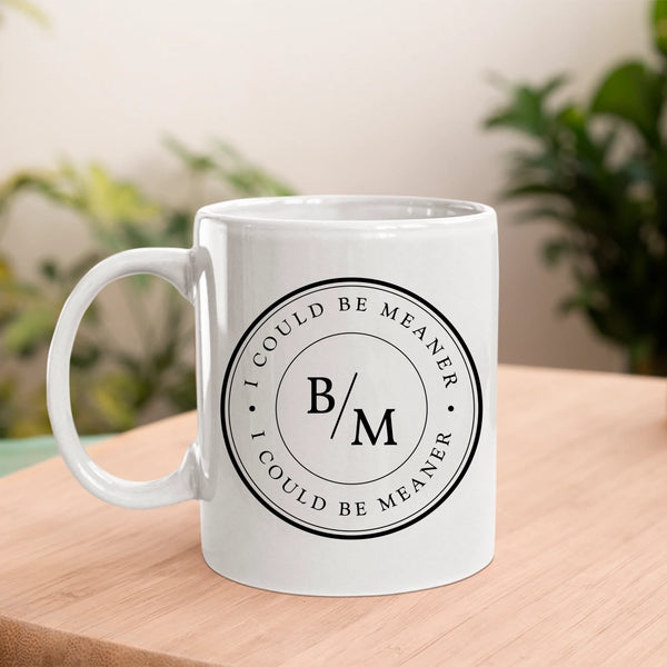 May include a white ceramic coffee mug displayed on a wooden surface, featuring a circular black design with the text “I COULD BE MEANER” repeated around the outer ring and the initials “B/M” centered inside. The background shows blurred green plants and soft natural light.