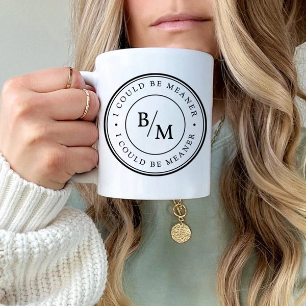 May include a white ceramic coffee mug displayed on a wooden surface, featuring a circular black design with the text “I COULD BE MEANER” repeated around the outer ring and the initials “B/M” centered inside. The background shows blurred green plants and soft natural light.