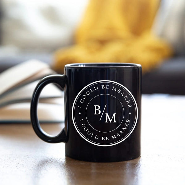 May include a white ceramic coffee mug displayed on a wooden surface, featuring a circular black design with the text “I COULD BE MEANER” repeated around the outer ring and the initials “B/M” centered inside. The background shows blurred green plants and soft natural light.