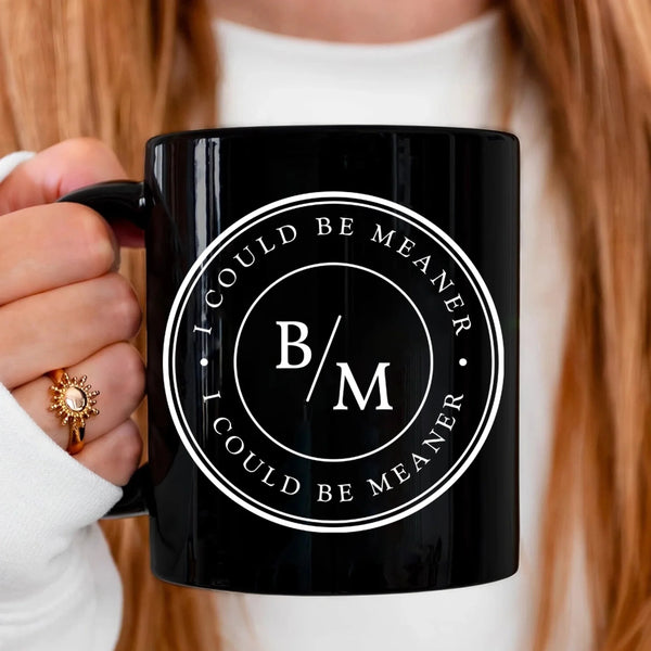 May include a white ceramic coffee mug displayed on a wooden surface, featuring a circular black design with the text “I COULD BE MEANER” repeated around the outer ring and the initials “B/M” centered inside. The background shows blurred green plants and soft natural light.