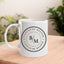 May include a white ceramic coffee mug displayed on a wooden surface, featuring a circular black design with the text “I COULD BE MEANER” repeated around the outer ring and the initials “B/M” centered inside. The background shows blurred green plants and soft natural light.