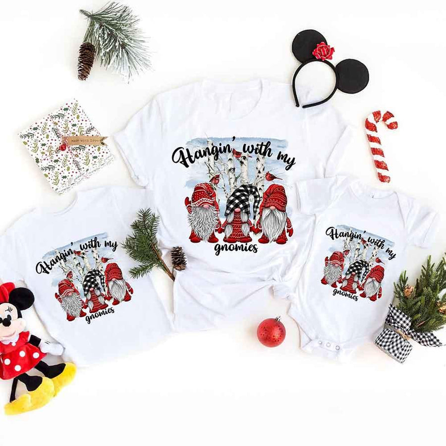 White t-shirt with Christmas-themed design featuring gnomes and text, surrounded by festive items on a white background.