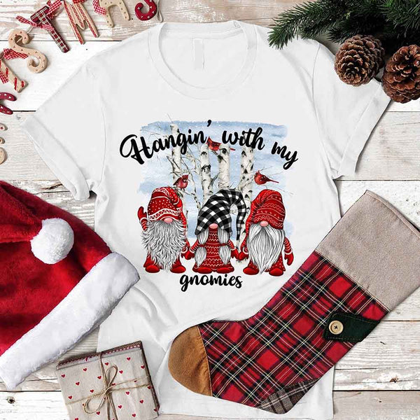 White t-shirt with Christmas-themed design featuring gnomes and text, placed on a wooden surface with festive items.