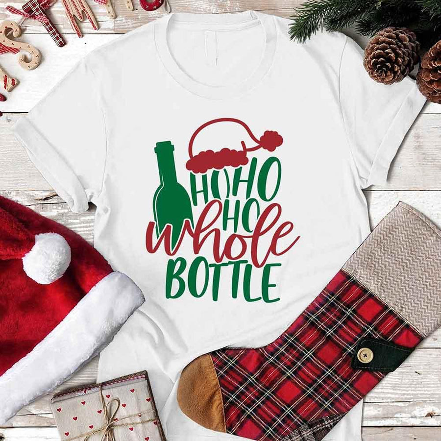White t-shirt with Christmas-themed design featuring a bottle and text, surrounded by festive items on a wooden surface.