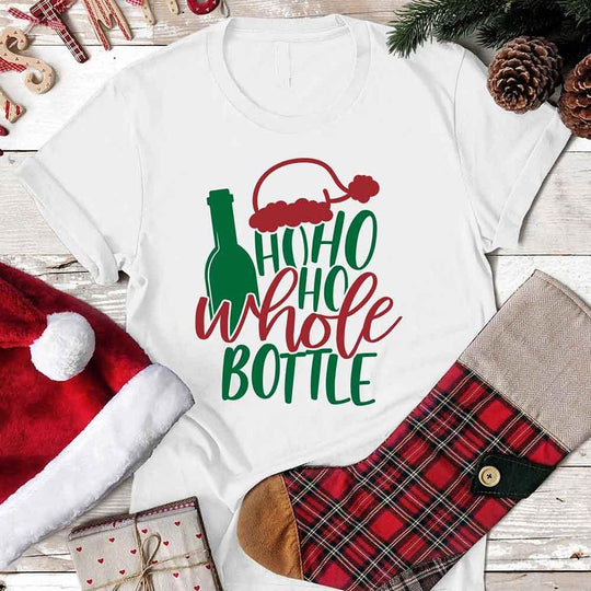 White t-shirt with Christmas-themed design featuring a bottle and text, surrounded by festive items on a wooden surface.