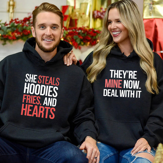 A man and a woman sitting together, wearing black hoodies with text on them. The man's hoodie reads 'She Steals Hoodies, Fries, and Hearts, They're Mine Now.