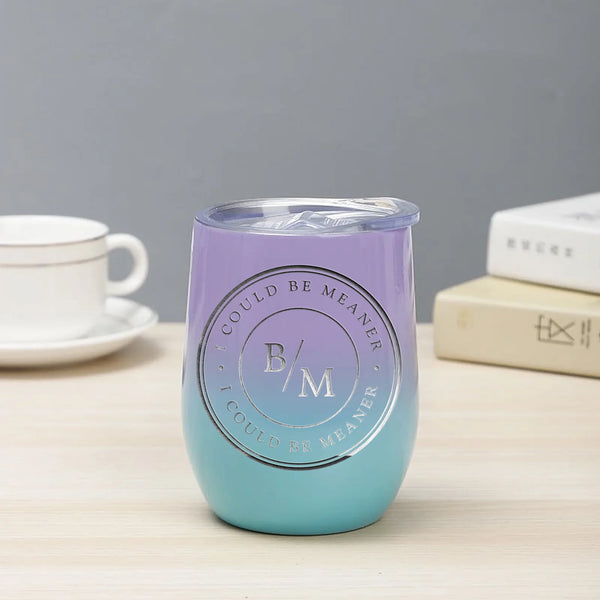 May include a wine tumbler displayed on a wooden surface, featuring a circular black design with the text “I COULD BE MEANER” repeated around the outer ring and the initials “B/M” centered inside. 