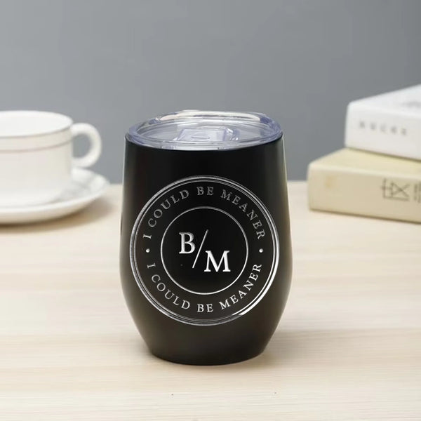 May include a wine tumbler displayed on a wooden surface, featuring a circular black design with the text “I COULD BE MEANER” repeated around the outer ring and the initials “B/M” centered inside. 