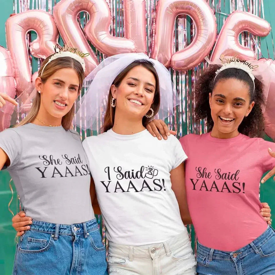 Three women wearing 'She Said YAAAS!' t-shirts with pink 'PRIDE' balloons in the background.