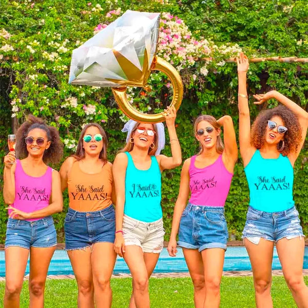 Five women in colorful tank tops and shorts celebrating outdoors with a large diamond-shaped balloon.