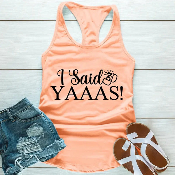 A Peach tank top with the text 'I Said YAAAS!' printed in black, placed on a wooden surface with blue denim shorts and white sandals beside it.