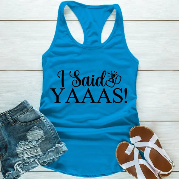 A Blue tank top with the text 'I Said YAAAS!' printed in black, placed on a wooden surface with blue denim shorts and white sandals beside it.